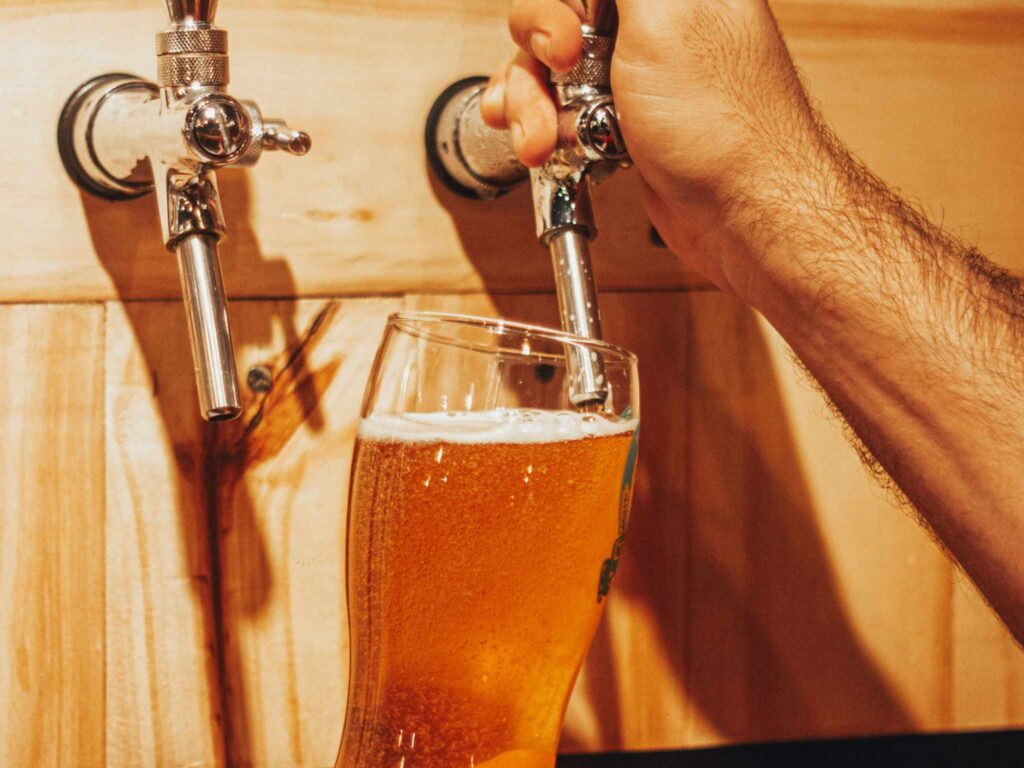 A person pours golden beer from a metal tap into a tilted glass.