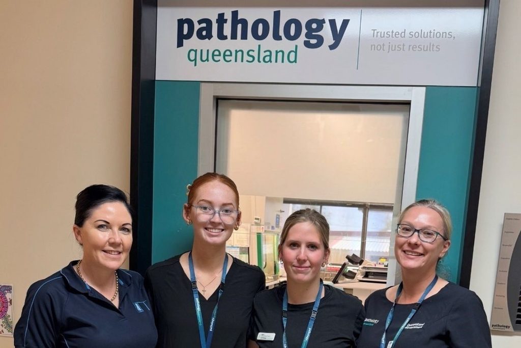 Four women, one wearing an LTT polo shirt, stand together beneath a Pathology Queensland sign.